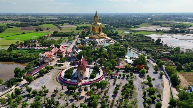 Aerial view dolly in establishing the giant Golden Buddha and the grand Wat Muang temple in Ang Thong, Thailand. A tourist area surrounded by rice paddies on a sunny day