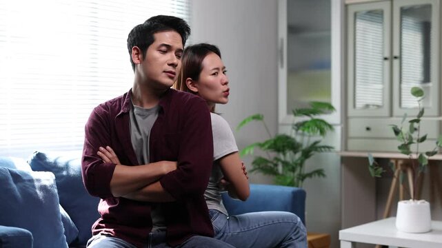 An Asian couple sits back to back on a living room couch, experiencing relationship tensions after an argument.