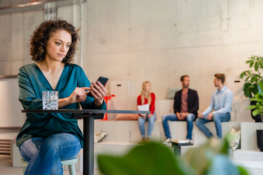 Businesswoman using mobile phone at desk with coworkers in background