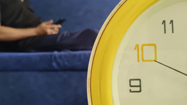 Clock ticking seconds needle in foreground and blurry background with lazy smartphone addict person lying on sofa doom scrolling on mobile phone. People wasting time on social media content concept.