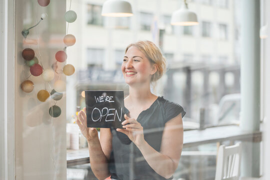 Smiling woman in a cafe attaching open sign to glass pane