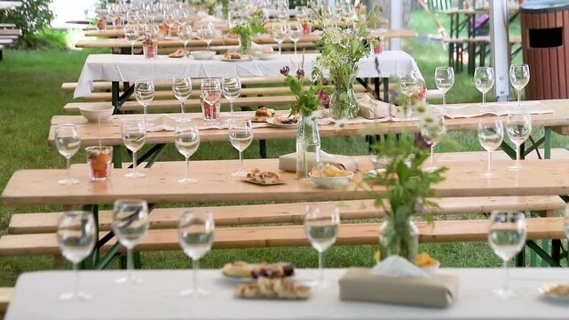 Eye-level shot of decorated wooden beer garden tables set up outdoors under a tent for a celebration, featuring wine glasses, wildflowers, and snacks.