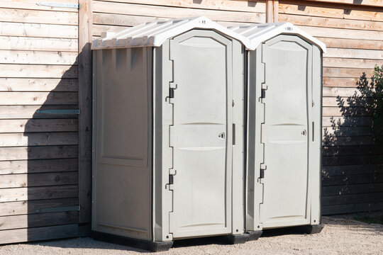 two portable plastic toilets standing side by side against a wooden wall in an outdoor area, concept of sanitation services for public events, construction site facilities, outdoor hygiene solutions