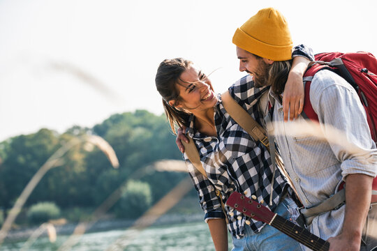 Happy young couple with backpacks and guitar embracing at the riverside