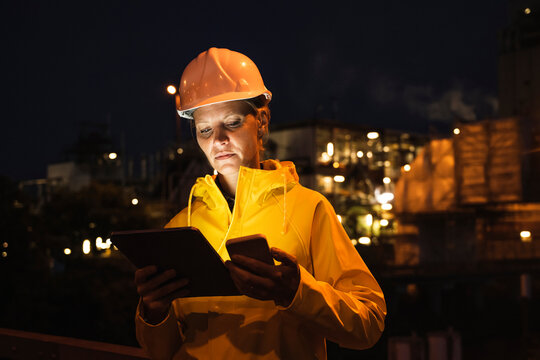 Female professional in yellow hardhat and raincoat using wireless technologies at night