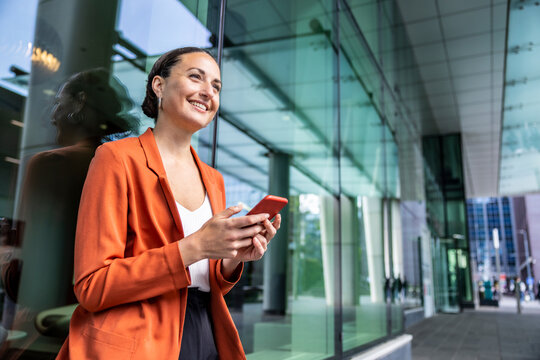 Happy businesswoman holding mobile phone leaning on glass wall
