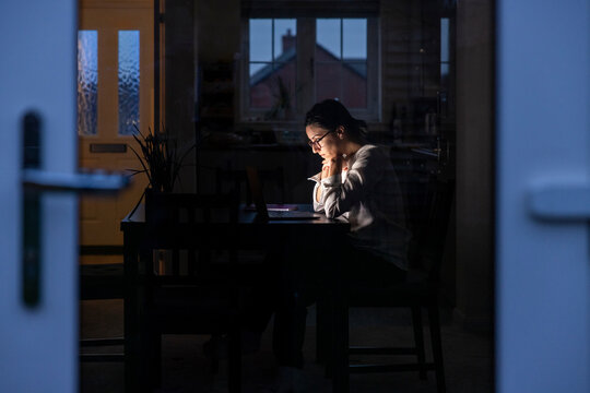 Thoughtful businesswoman looking at laptop while working during night seen through glass door