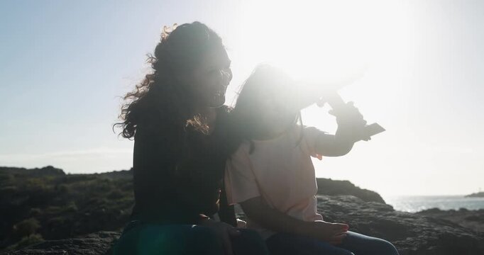 Happy latin mother having playful time with daughter using wood airplane on the beach during sunset - Holiday, summer and family lifestyle concept