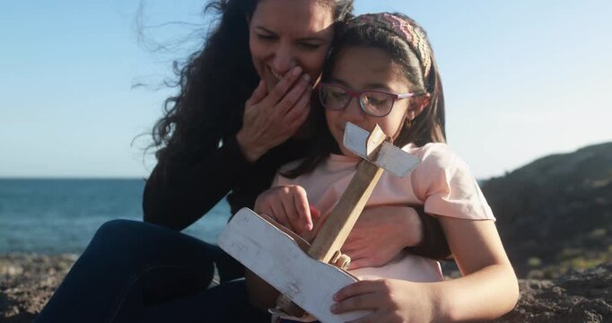 Happy latin mother having playful time with daughter using wood airplane on the beach - Holiday, mom day and family lifestyle concept