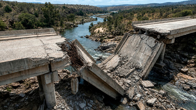 Collapsed concrete bridge over a river. Destroyed infrastructure with exposed steel rebar and rubble. Natural disaster damage concept
