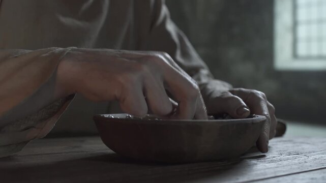 Close up of medieval prisoner hands scraping porridge with a slice of dark bread from a wooden bowl on a rustic table inside a gloomy dungeon