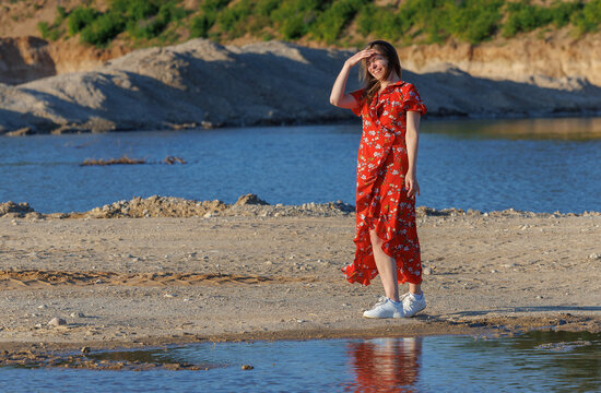 Young woman in red summer dress walking by blue lake on a sunny day