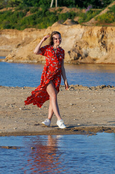 Young woman in red summer dress walking by blue lake on a sunny day