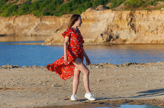 Young woman in red summer dress walking by blue lake on a sunny day