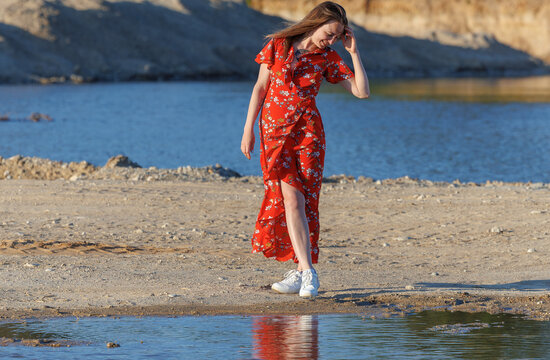 Young woman in red summer dress walking by blue lake on a sunny day