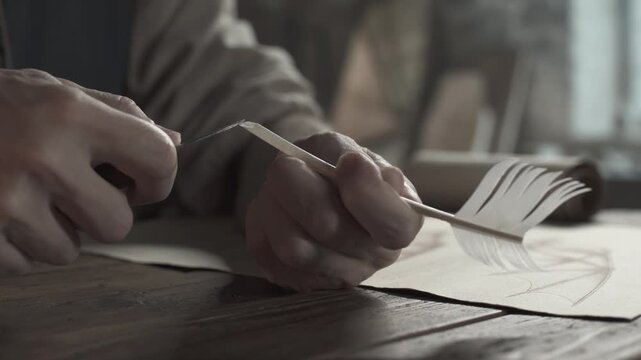 Close up of medieval scholar hands cutting the tip of a quill pen with a knife and checking it with a thumb next to blueprint on a rustic wooden desk inside a dimly lit workshop