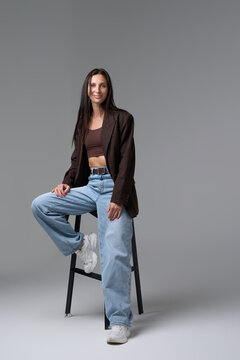 Fashionable woman in brown blazer and blue jeans sitting on high chair in studio