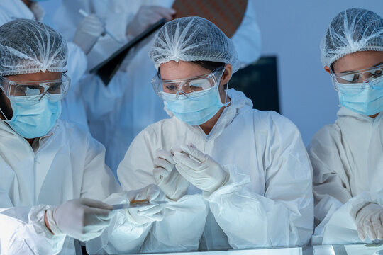 Three specialists in white protective suits and masks studying small samples on an illuminated table, semiconductor inspection in cleanroom facility.