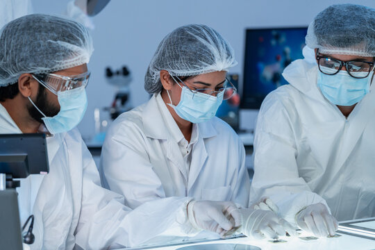 Three specialists in protective suits and masks examining material specimens on an illuminated table, semiconductor production in blue-lit cleanroom.