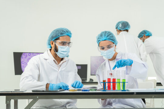 Scientists in masks and goggles examining a test tube with sample, rack of red and green tubes on bench, research discussion in bright lab.