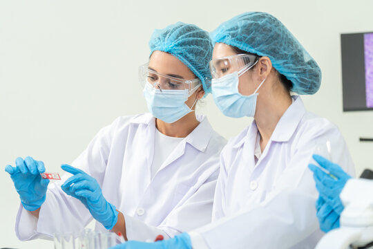 Women in masks and goggles inspecting a red specimen slide together at the bench, focused medical sample analysis in a bright research lab.