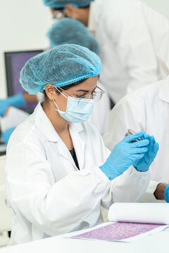 Young woman in glasses and hairnet inspecting a sample in gloved hands, histology printout on the bench, focused research work in bright lab.