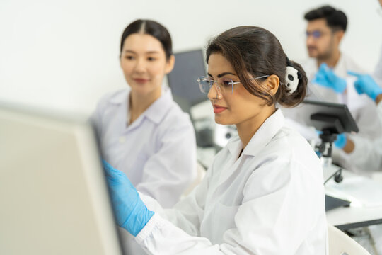 Asian woman with glasses focused on a screen while smiling colleague points, male researcher behind, friendly research teamwork in bright lab.