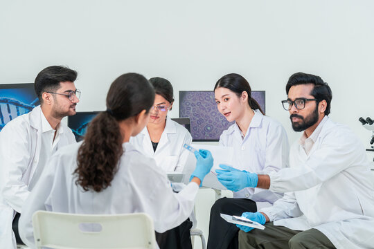 Five scientists in lab coats talking around a test tube, DNA image on screen behind, collaborative team discussion in bright research laboratory.