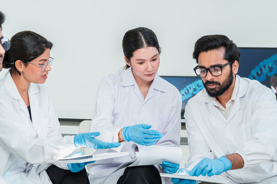 Two men and a woman in lab coats and gloves studying paperwork during a discussion, science team collaboration in a bright research laboratory.