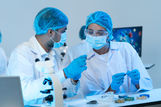 Man and woman in masks and goggles examining tiny samples at a bench with microscope and petri dishes of material, research teamwork in blue light.