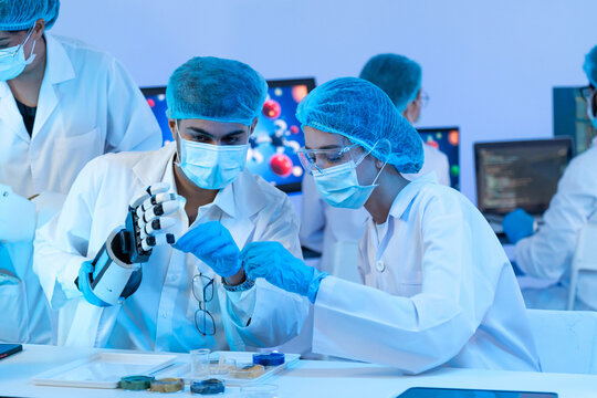Man and woman in masks and hairnets inspecting a black robotic prosthetic hand at the bench, robotics research teamwork in blue-lit laboratory.