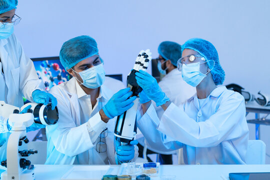 Man and woman in masks, hairnets and goggles examining a white robotic hand and arm under blue light, robotics and prosthetic research teamwork.