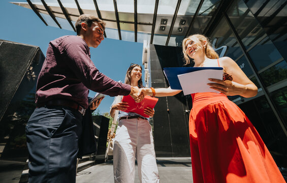Business colleagues shake hands outside a modern office building while exchanging documents and smiling. A man and two women meet at an office entrance in a friendly professional greeting.