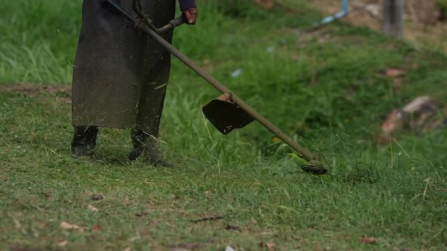 Grass lawn weed trimmer string trimmer gardener maintenance cutting outdoor vegetation close up trimming green field, focused yard care with brushcutter, apron boots, flying clippings debris