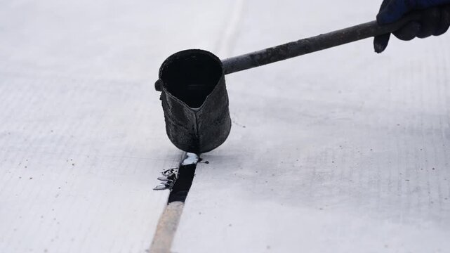 Asphalt crack repair sealant for pavement road maintenance, construction worker pouring hot bitumen with ladle into joint, roof seam waterproofing tar application on flat surface to prevent leaks