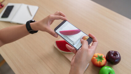 Naklejka premium Cropped hands holding smartphone to take photo of toy watermelon on table