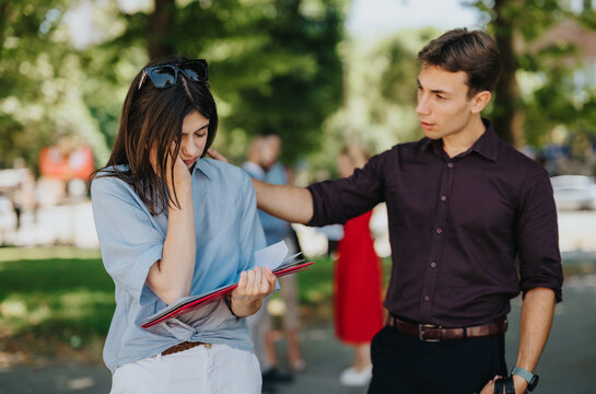 A young man gently comforts an upset woman who is holding documents outdoors. The couple shares a quiet moment of support and concern in a park setting.