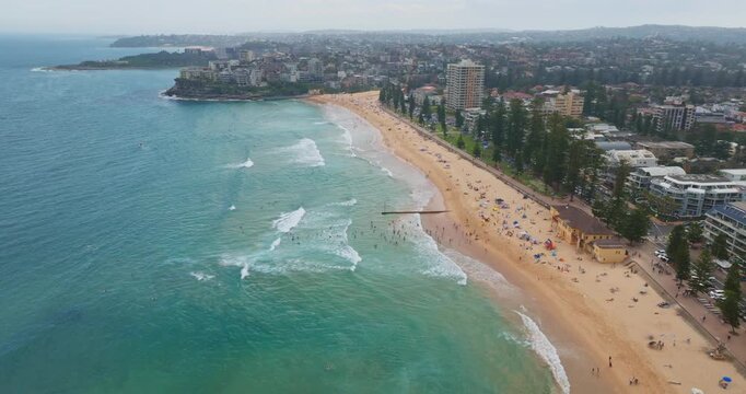 Expansive Manly Beach in Sydney, Australia, showing the vibrant coastline with people relaxing, swimming, and surfing in the clear turquoise ocean waters under a summer sky. Aerial view drone flight