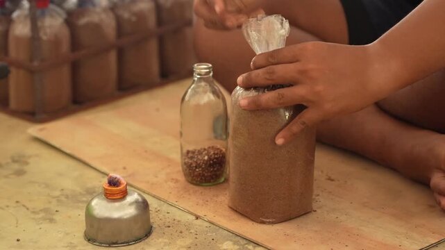 Hand craft workshop sealing homemade process adult hands fill plastic bag and glass bottle with sand using funnel, alcohol lamp burner flame heating nearby, DIY recycling indoor workspace closeup