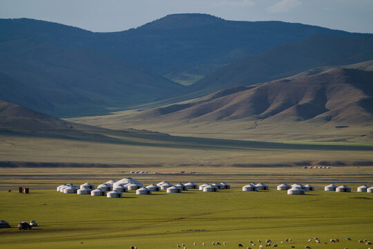 Vast Mongolian steppe with yurts scattered across the green plains, set against a backdrop of rolling mountains under a clear blue sky.