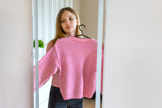 Young Woman Holding Pink Sweater in Front of a Mirror. Young woman holding a pink knitted sweater on a hanger while viewing it in a full-length mirror indoors.