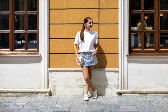 Young Woman Standing Against a Sunlit Yellow Building. Young woman in casual summer clothes standing against a sunlit yellow building between two large wooden windows.