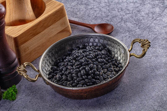 Black beans in hammered metal bowl with wooden spice mill and spoon on textured gray surface