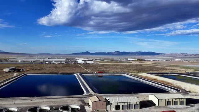 Aerial footage of water reservoirs and a hyperscale data center in Utah's West Desert, backdropped by a dramatic mountain range and dynamic sky, in bright midday light.