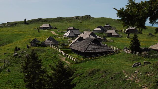Traditional wooden huts sit across a green alpine plateau with fenced pastures, scattered rocks and rolling hills under bright summer daylight.