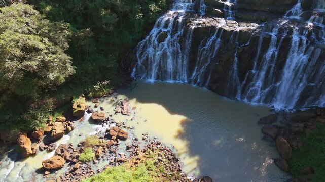 Aerial drone view reveals the full width of Salto Yasi waterfall cascading into an ochre pool.