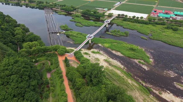 Aerial tilt up glides from Yeoman-ri Pedestrian Bridge skywalk over Pyeongchang River wetlands toward farmland in mountain valley