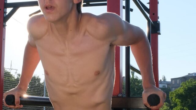 Slim young man with a muscular body performs dips on parallel bars at sunrise. Shirtless athlete training alone outdoors, calisthenics, natural fitness, bodyweight training, weight management.