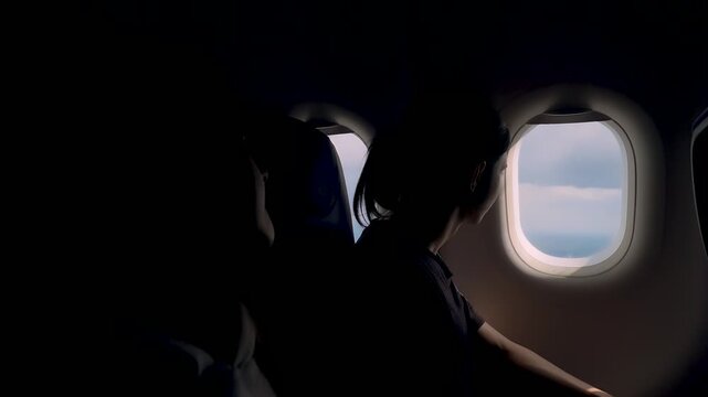Silhouetted teenagers looking at window view during airplane trip high above earth. Brother and sister sitting side by side inside aircraft cabin. Flight travel concept freedom calm scenery