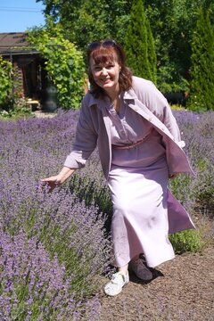 Woman in a lavender field bends down to admire the flowers, dressed in a pastel dress with a cheerful smile, surrounded by lush greenery on a sunny day in a serene garden